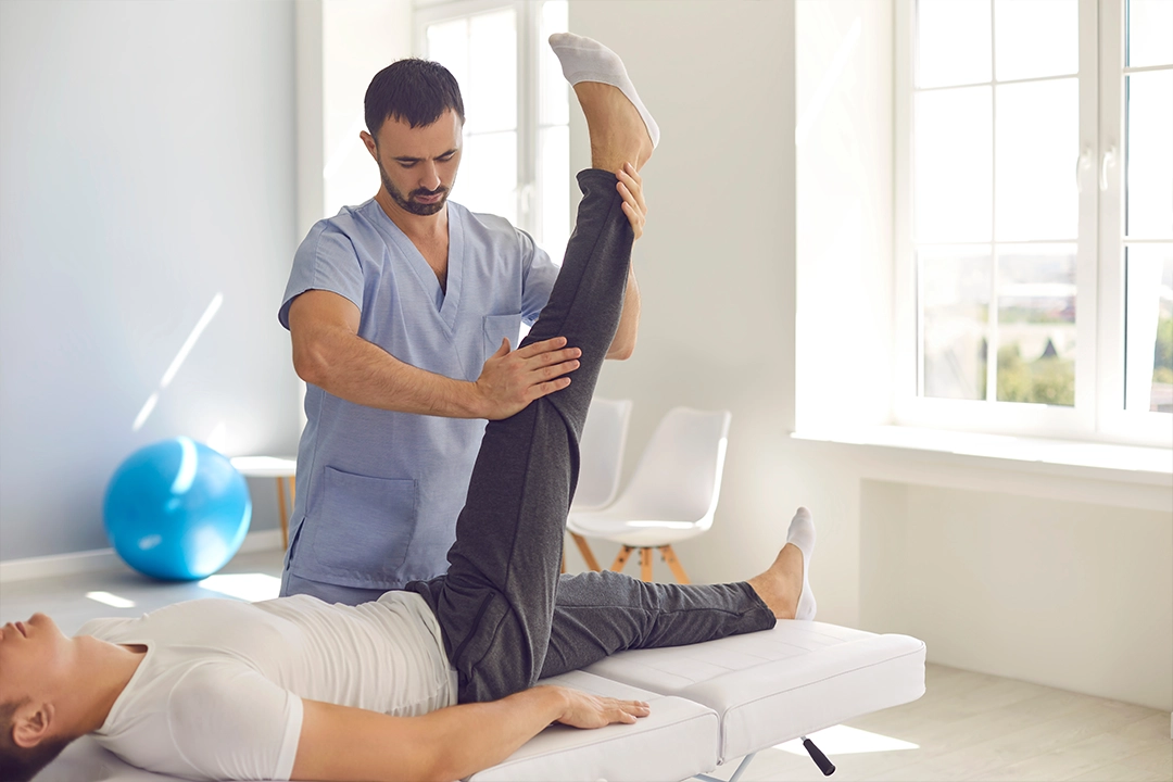 A physical therapist assists a patient by stretching their leg on a treatment table in a bright, modern therapy room.