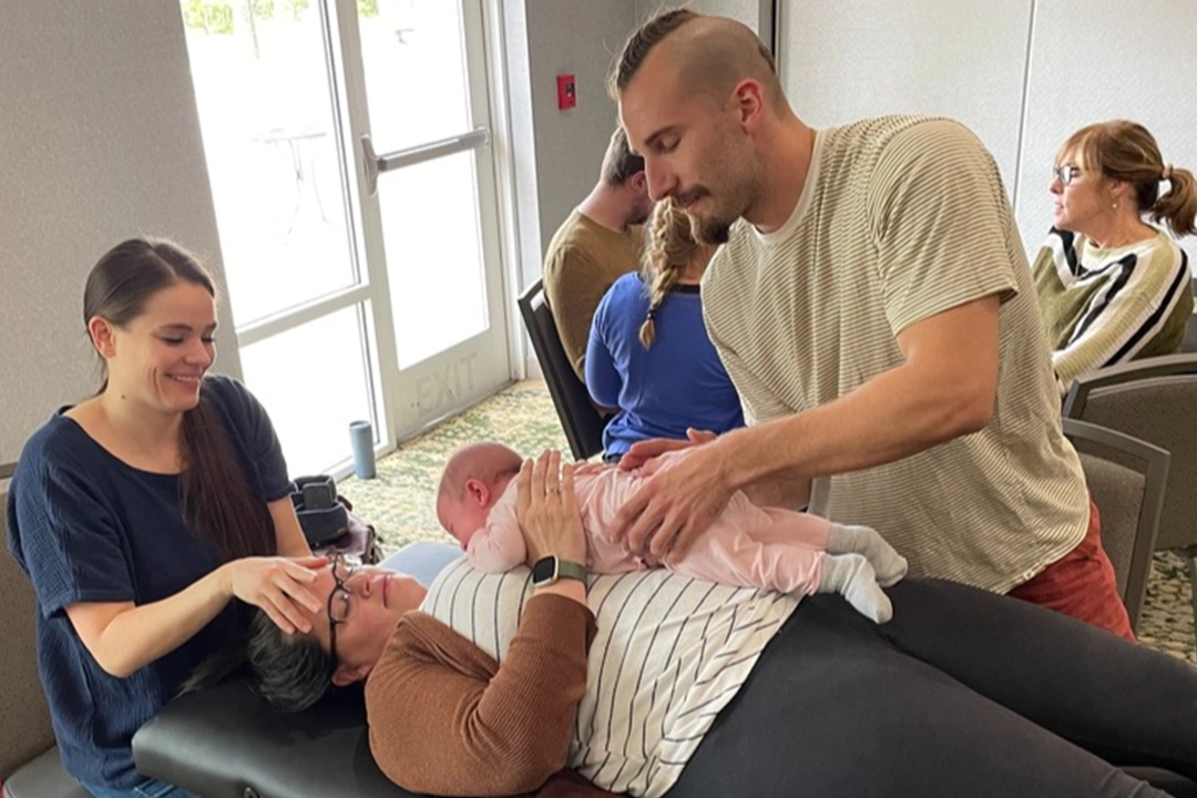 A man tenderly holds a baby on a woman's lap as another woman massages the mother's head in a relaxed indoor setting.